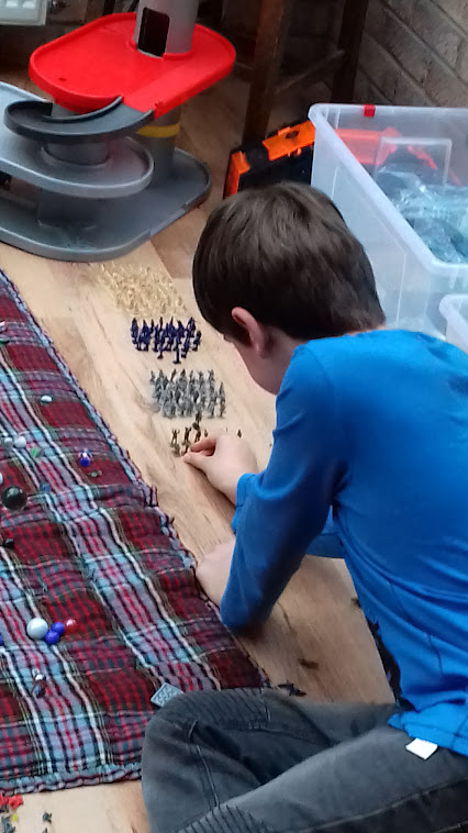 A boy, seen from behind, kneeling on a wooden floor at home carefully arranging rows of toy soldiers
